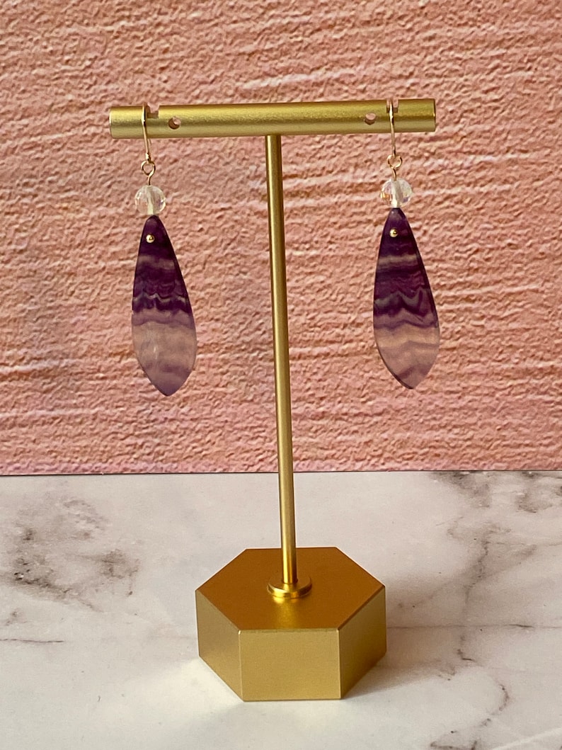 Pode incluir: Um par de brincos pendentes com pedras em forma de l&aacute;grima listradas de roxo e branco e contas de cristal transparentes penduradas em ganchos dourados.