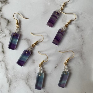May include: A pair of earrings with purple and blue fluorite stones and gold hooks. The stones are rectangular and have a clear crystal bead above them.