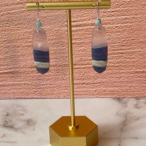 Pode incluir: Um par de brincos pendentes com uma pedra azul claro e roxa em cada brinco. Os brincos est&atilde;o pendurados em um suporte dourado.