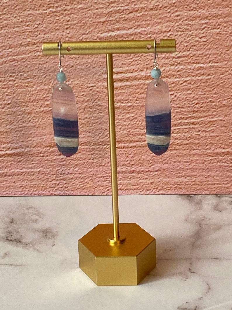 Pode incluir: Um par de brincos pendentes com uma pedra azul claro e roxa em cada brinco. Os brincos est&atilde;o pendurados em um suporte dourado.