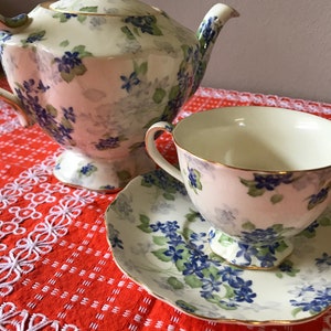 May include: A white teapot and teacup with a floral pattern of blue and green flowers. The teapot has a lid and a spout. The teacup has a handle and a saucer. The teapot and teacup are on a red and white tablecloth.