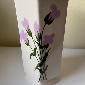 May include: White ceramic vase with a geometric shape and a floral design featuring pink flowers and green stems.