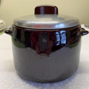 May include: A dark brown ceramic pot with a silver lid and a brown handle. The pot has two small handles on the sides. The pot is sitting on a white surface with small white polka dots.