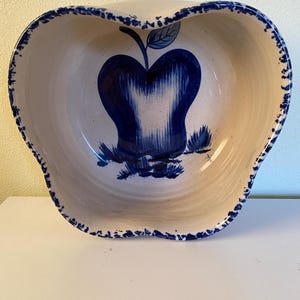 May include: Cream-colored ceramic apple-shaped bowl with a blue apple design in the center. The bowl has a scalloped edge with a blue dotted trim. The apple design features a blue stem and leaf.