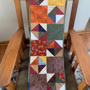 May include: A handmade patchwork quilt runner with a variety of fabrics in shades of orange, brown, green, and yellow. The quilt is placed on a wooden chair. The runner features a repeating square pattern with geometric designs.