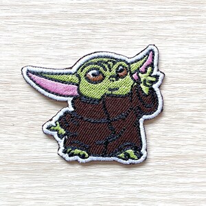 May include: An iron-on patch embroidered with a green cartoon character with large ears, wearing a brown robe. The character has a mischievous expression and is holding a finger to its mouth.