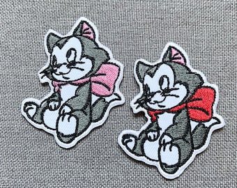 May include: Two black and white embroidered iron-on patches of cartoon cats with pink and red bows. The cats are sitting with their tails curled around their legs.