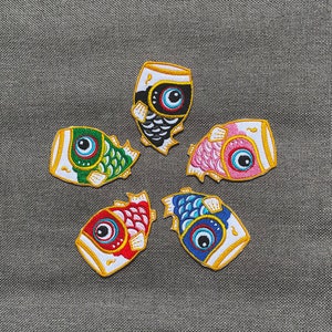 May include: Six embroidered fish-shaped patches in various colors, including black, green, pink, blue, and red. Each patch features a stylized fish with a large eye and a gold outline.