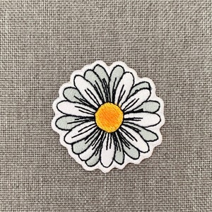 May include: A white daisy embroidered patch with a yellow center. The patch is on a gray fabric background.