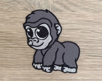 Può includere: Un toppa ricamata grigia di un gorilla dei cartoni animati con occhi grandi. Il gorilla è in piedi a quattro zampe e ha un'espressione amichevole.