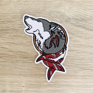 Könnte beinhalten: Gestickter Bügel-Patch mit einem heulenden grauen Wolf in einem Traumfänger mit roten und braunen Federn.