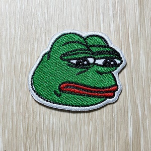 Könnte beinhalten: Grüner gestickter Aufnäher eines traurigen Frosch-Memes mit rotem Mund und weißen Augen.