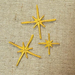 Könnte beinhalten: Drei goldene Sternen-Verzierungen in verschiedenen Größen. Die Sternen haben mehrere spitze Strahlen, die sich von einem zentralen Punkt aus erstrecken. Die Sterne sind auf einer strukturierten, neutralfarbenen Oberfläche angeordnet.