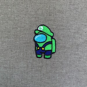 May include: Embroidered patch featuring a green Among Us character dressed as Luigi, with a green hat and blue overalls. The character has a blue visor and is outlined in black. The patch is on a gray fabric background.