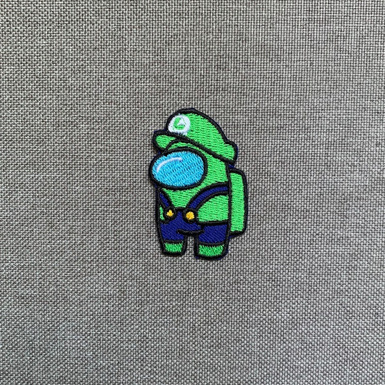 May include: Embroidered patch featuring a green Among Us character dressed as Luigi, with a green hat and blue overalls. The character has a blue visor and is outlined in black. The patch is on a gray fabric background.