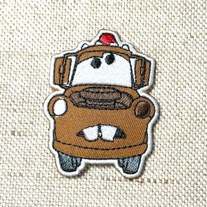 Könnte beinhalten: Gestickter Aufnäher mit einer Zeichentrickfigur eines Abschleppwagens in Braun, Weiß und Schwarz. Der Aufnäher hat ein rotes Licht oben und ist weiß umrandet. Das Design befindet sich auf einem beigefarbenen Stoffhintergrund.