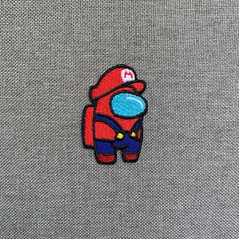 May include: Embroidered patch featuring a red character with a blue visor and a red hat with a white "M" symbol. The character is wearing blue overalls with yellow buttons. The patch is on a gray fabric background.