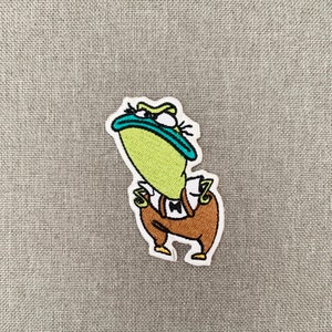 May include: An embroidered iron-on patch featuring a green cartoon frog wearing brown trousers and a bow tie. The frog has a grumpy expression and is looking to the left.