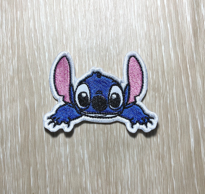 May include: Blue and pink embroidered character with large eyes and a wide smile. The character is stitched in a cartoon style and has a white outline.