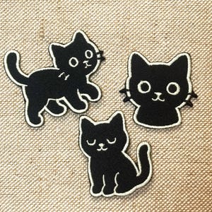 May include: Three black cat patches with white embroidered outlines. One cat is walking, one is a cat face, and the third is sitting. The patches are on a tan fabric background.