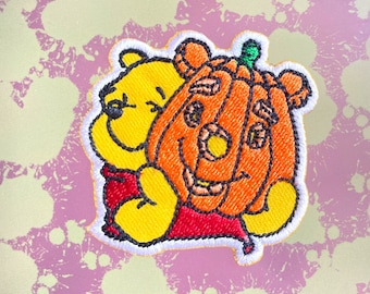 May include: Embroidered patch featuring Winnie the Pooh holding a smiling jack-o'-lantern pumpkin. The patch is yellow, orange, red, and white.