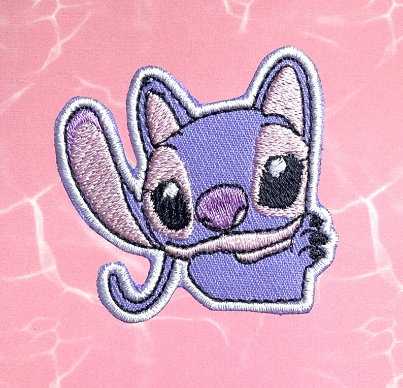 May include: Purple and white embroidered iron-on patch of Stitch from Lilo & Stitch, with a white outline and a pink nose.