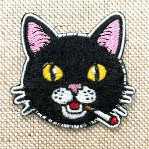 Pode incluir: Patch bordado com a cabeça de um gato preto com olhos amarelos, orelhas cor-de-rosa e língua vermelha. O gato está segurando um cigarro na boca. O patch tem uma borda branca e está em um fundo de tecido bege.