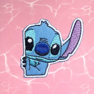May include: Blue and purple embroidered iron-on patch of a cartoon character with big eyes and a single ear. The character is Stitch from the Disney movie Lilo & Stitch.