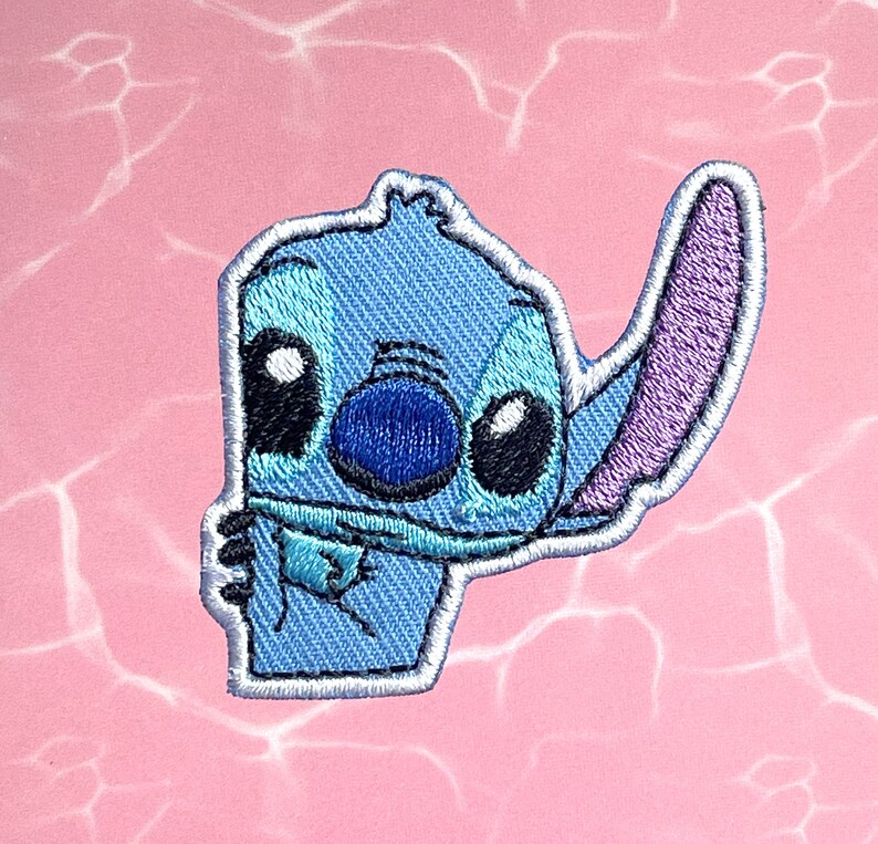 May include: Blue and purple embroidered iron-on patch of a cartoon character with big eyes and a single ear. The character is Stitch from the Disney movie Lilo & Stitch.