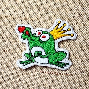 Może przedstawiać: Haftowana naszywka przedstawiająca zieloną żabę w złotej koronie trzymającą czerwone serce. Żaba ma duże białe oczy z czerwonymi źrenicami i jest obrysowana na biało. Naszywka znajduje się na beżowym tle z tkaniny.