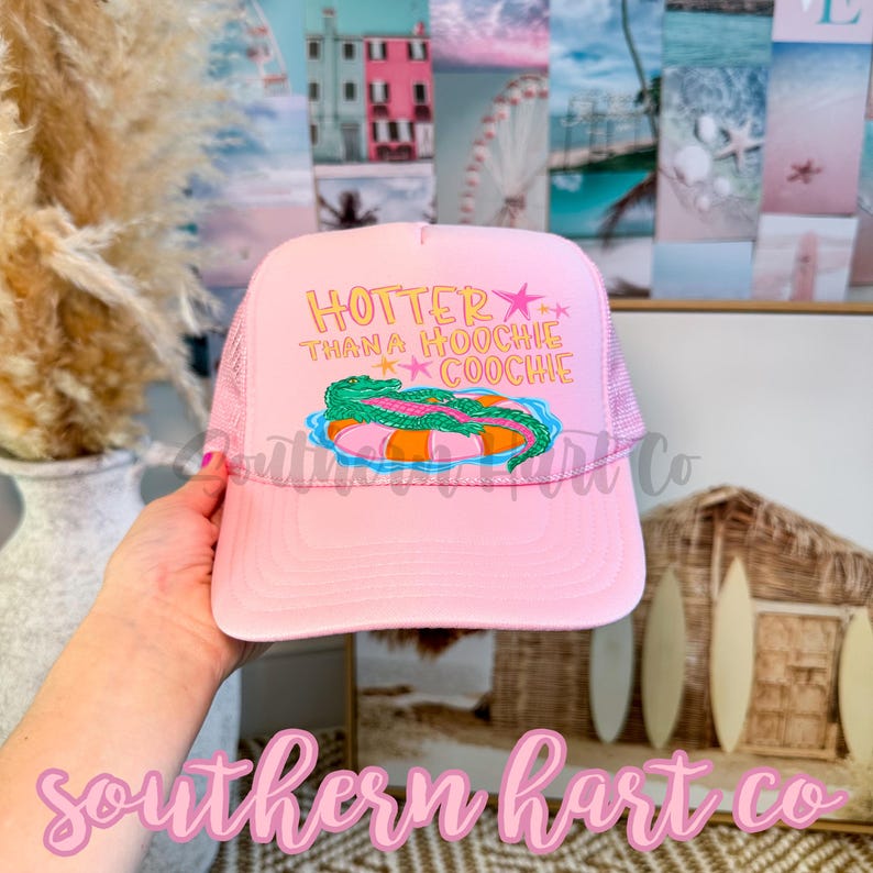 May include: Pink trucker hat with the text "Hotter Than a Hoochie Coochie" in yellow and a graphic of an alligator on an inner tube. The hat has a mesh back.