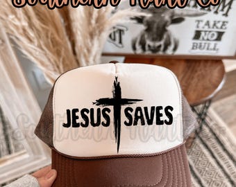 May include: A brown and white trucker hat with the text "JESUS SAVES" and a black cross graphic. The hat has a brown brim and mesh back. The brand name "Southern Hart Co" is above the hat.