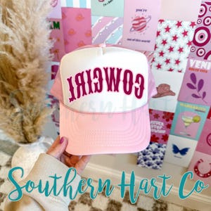 May include: Pink trucker hat with a white front panel. The word "Cowgirl" is printed in a dark pink, curved font. The hat has a mesh back and a curved brim. The hat is held up by a person.