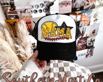 May include: A black and white trucker hat with a white mesh back. The front of the hat features a yellow softball with red stitching, a black star with glitter, and the word "MAMA" in yellow letters with red accents.