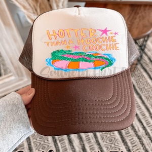 May include: A brown and white trucker hat with the text "HOTTER THAN A HOOCHIE COOCHIE" in pink and yellow. The hat features a graphic of an alligator on an inner tube. The brim is brown, and the mesh is a light brown color.