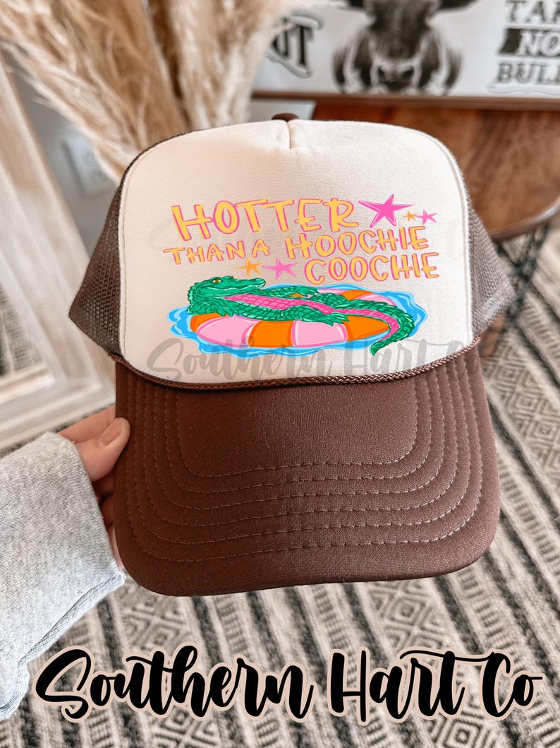 May include: A brown and white trucker hat with the text "HOTTER THAN A HOOCHIE COOCHIE" in pink and yellow. The hat features a graphic of an alligator on an inner tube. The brim is brown, and the mesh is a light brown color.