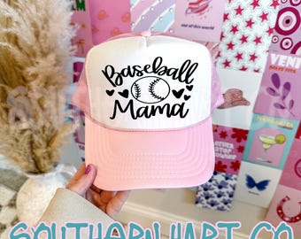 May include: Pink and white baseball cap with the text "Baseball Mama" in black script, featuring a baseball graphic and heart accents. The cap has a mesh back and a curved brim.