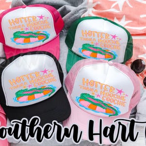 May include: Four trucker hats in pink, black, green, and light pink. Each hat has a white front panel with the text "HOTTER THAN A HOOCHIE COOCHIE" and a graphic of a pool float. The hats are on a patterned background.
