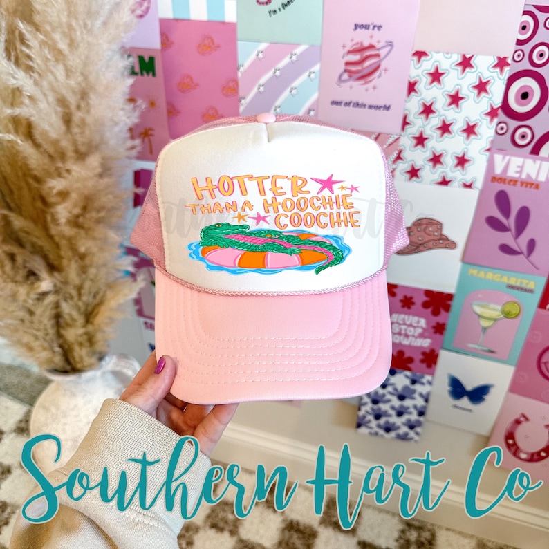 May include: Pink trucker hat with a white front panel featuring the text "HOTTER THAN A HOOCHIE COOCHIE" above an illustration of a pool float. The hat has a pink brim and mesh back.