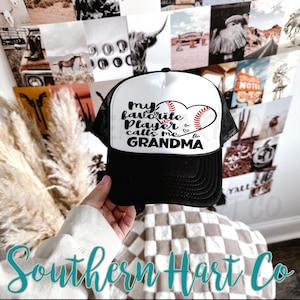 May include: Black and white baseball cap with the text "my favorite player calls me GRANDMA" in black script. The design includes a baseball heart. The cap has a mesh back and a curved brim.