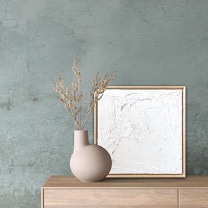 May include: A white textured canvas painting in a light wood frame sits on a light wood dresser with a beige ceramic vase with dried beige branches.