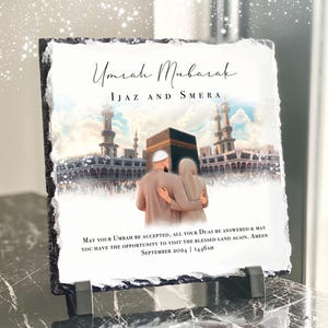 May include: A black slate plaque with a white background and a picture of a couple standing in front of the Kaaba in Mecca. The text "Umrah Mubarak" is written in a decorative font at the top of the plaque. The text "IJAZ AND SMERA" is written below the title. The text "MAY YOUR UMRAH BE ACCEPTED, ALL YOUR DUAS BE ANSWERED & MAY YOU HAVE THE OPPORTUNITY TO VISIT THE BLESSED LAND AGAIN. AMEEN SEPTEMBER 2024 | 1446AH" is written at the bottom of the plaque.