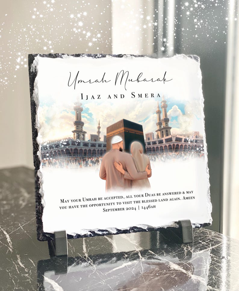 May include: A black slate plaque with a white background and a picture of a couple standing in front of the Kaaba in Mecca. The text "Umrah Mubarak" is written in a decorative font at the top of the plaque. The text "IJAZ AND SMERA" is written below the title. The text "MAY YOUR UMRAH BE ACCEPTED, ALL YOUR DUAS BE ANSWERED & MAY YOU HAVE THE OPPORTUNITY TO VISIT THE BLESSED LAND AGAIN. AMEEN SEPTEMBER 2024 | 1446AH" is written at the bottom of the plaque.