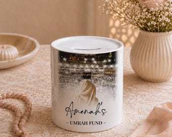 May include: A white ceramic money box featuring a photographic print of the Kaaba in Mecca with a person in a white garment. The text reads Amenah's - UMRAH FUND - in black script and block lettering. The cylindrical container has a coin slot on top.