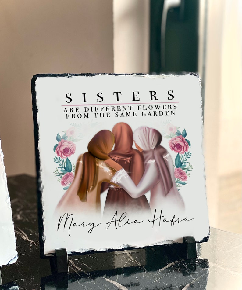 Puede incluir: Una placa de pizarra blanca con borde negro que presenta una cita sobre las hermanas que son flores diferentes del mismo jard&iacute;n. La cita est&aacute; rodeada de flores rosas y hojas verdes. Tres mujeres con pa&ntilde;uelos en la cabeza est&aacute;n de pie juntas en el centro de la placa. Los nombres Mary, Alia y Hafsa est&aacute;n escritos en cursiva en la parte inferior de la placa.