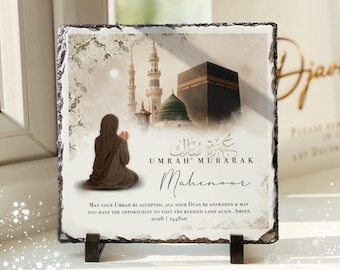 May include: A square slate plaque with rough edges featuring a printed image of the Kaaba and a person in prayer. The text reads: UMRAH MUBARAK, Mahenoor, and a prayer for acceptance of Umrah, dated 2026 | 1448AH. The plaque rests on two black stands.