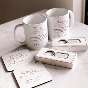 Könnte beinhalten: Zwei weiße Keramikbecher mit dem Text "Half Her Deen" in arabischer Schrift und Englisch. Die Becher sind mit einem goldfarbenen Design verziert. Der Text enthält auch die Namen "Zakariya" und "Sara" sowie das Datum "27.08.21". Zwei passende Untersetzer mit dem gleichen Text und Design sind ebenfalls abgebildet. Ein silberner Schlüsselanhänger mit dem gleichen Text und Design ist in einer weißen Schachtel abgebildet.
