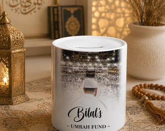 May include: A white ceramic money box featuring a printed image of a person in a white cap facing the Kaaba in Mecca. The text "Bilal's - UMRAH FUND -" is printed in black script and block lettering on the front of the cylindrical container.