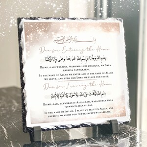 Könnte beinhalten: Eine Schiefertafel mit beigefarbenem Hintergrund und goldenen Glitzerakzenten. Die Tafel zeigt zwei islamische Gebete in arabischer Schrift mit englischen Übersetzungen darunter. Das erste Gebet ist für den Eintritt in das Haus, das zweite für das Verlassen des Hauses. Die Tafel ist ein dekoratives Stück für ein Zuhause.