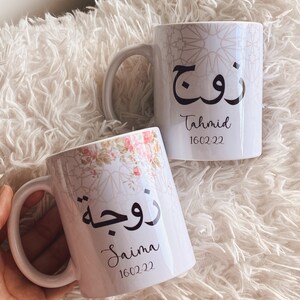 May include: Two white ceramic mugs with Arabic calligraphy and names. One mug features a floral design and the name "Saima" with the date 16.02.22. The other mug has the name "Tahmid" and the date 16.02.22.